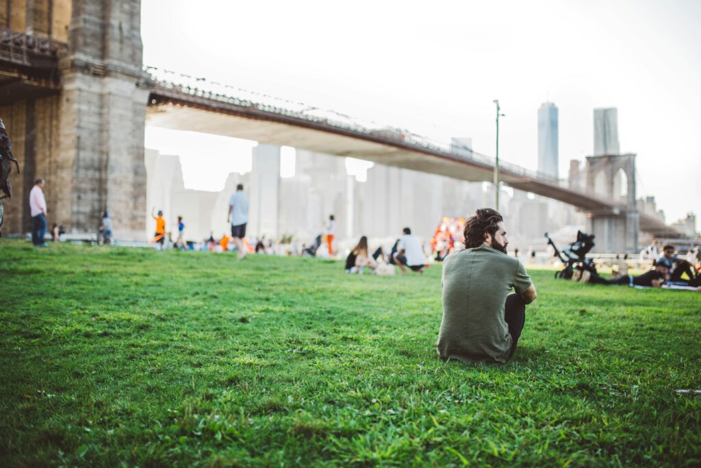 A young man sits on the grass by himself while people lounge along a riverbank in front of a bridge. It's easy to feel alone in the crowd if you don't know how to combat loneliness.